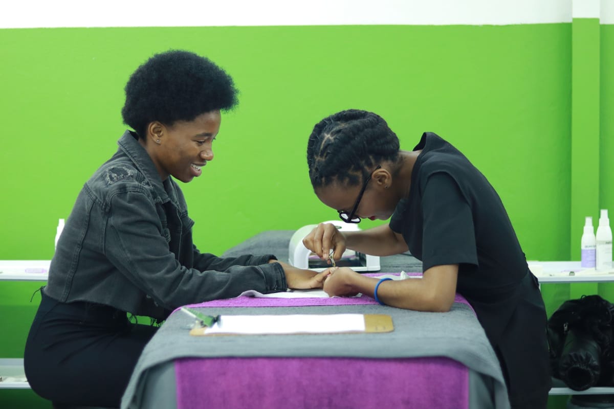 Two women sit across a table; one smiles whilst having her nails done by the other, who is focused on her work. The background features a bright green and white wall, and the table is covered with a grey cloth and a purple towel. Nail supplies are visible.