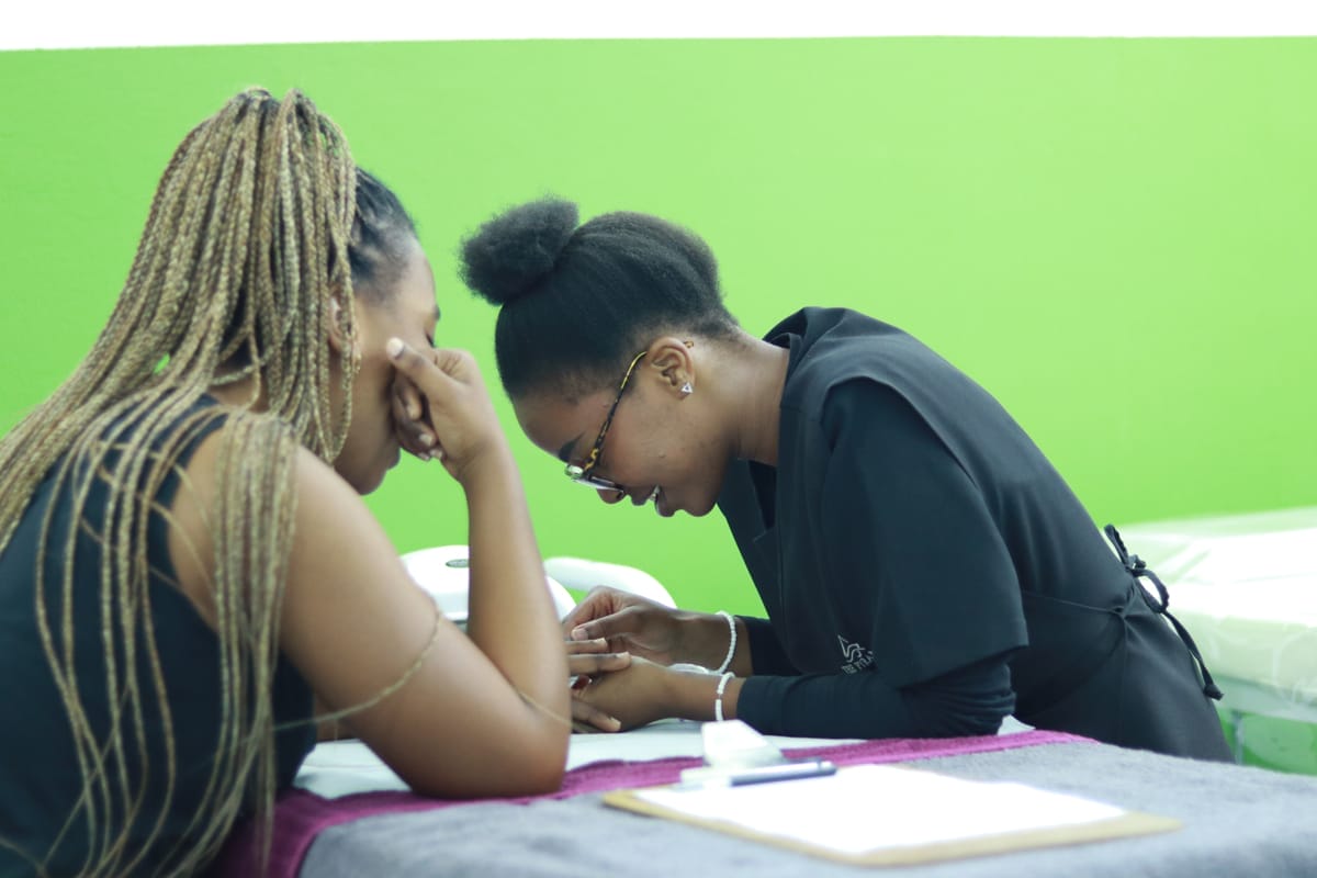 Two women sit opposite each other at a table covered with towels. One, with plaited hair, rests her chin on her hand whilst the other, wearing glasses and a black outfit, focuses intently as she gives a manicure. The background wall is bright green.