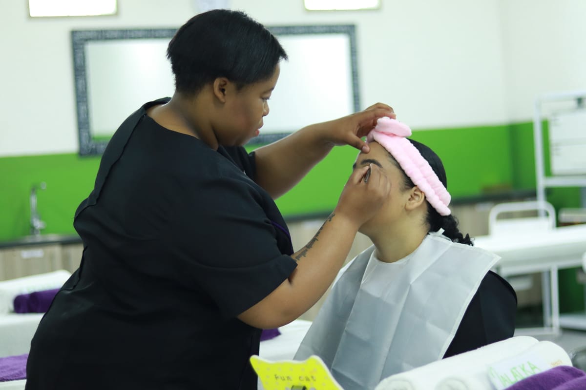 A beauty professional applies make-up to a seated client wearing a pink headband and a white protective cape in a well-lit, green and white salon with mirrors and treatment beds in the background.
