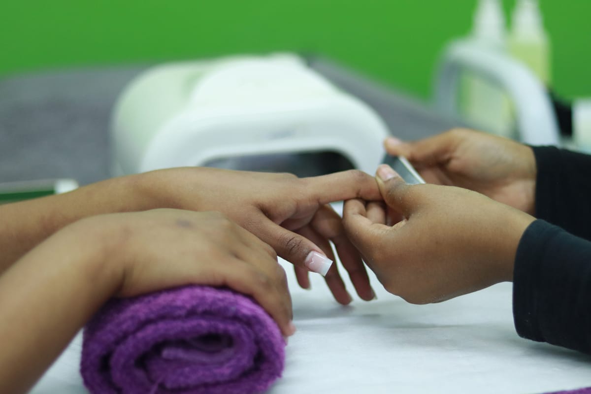 A person receives a manicure as another person files their fingernails. One hand rests on a purple towel, while nail care tools and bottles are visible in the background on a white surface. The scene is set against a green backdrop.