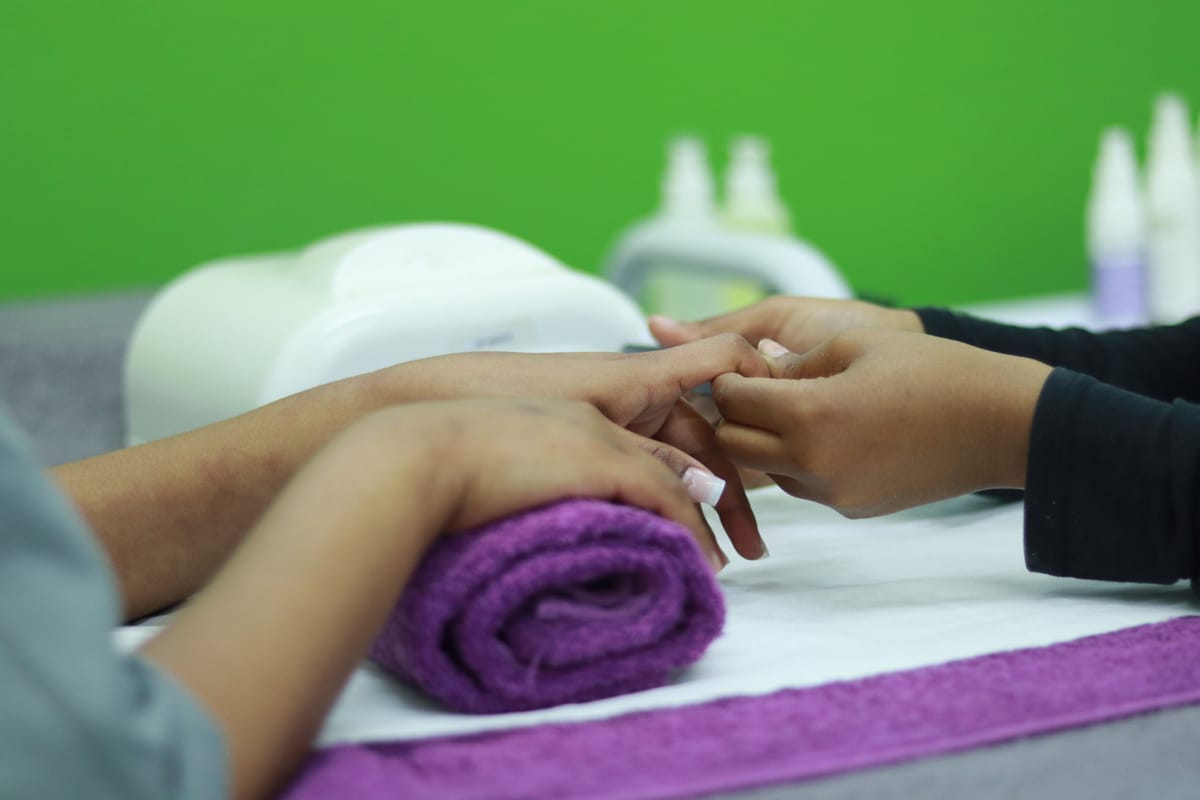 A person is receiving a manicure; one hand rests on a rolled purple towel whilst another person works on their nails. The background is green, and bottles of manicure products are visible on the table. The focus is on the hands and nail care process.