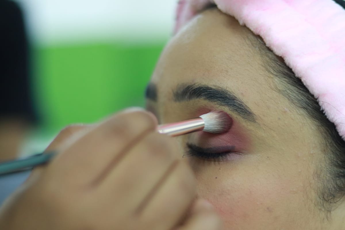Close-up of a person’s face with closed eyes as someone applies pink eyeshadow to their eyelid using a make-up brush. The person wears a soft pink hairband, and the background is blurred green and white.