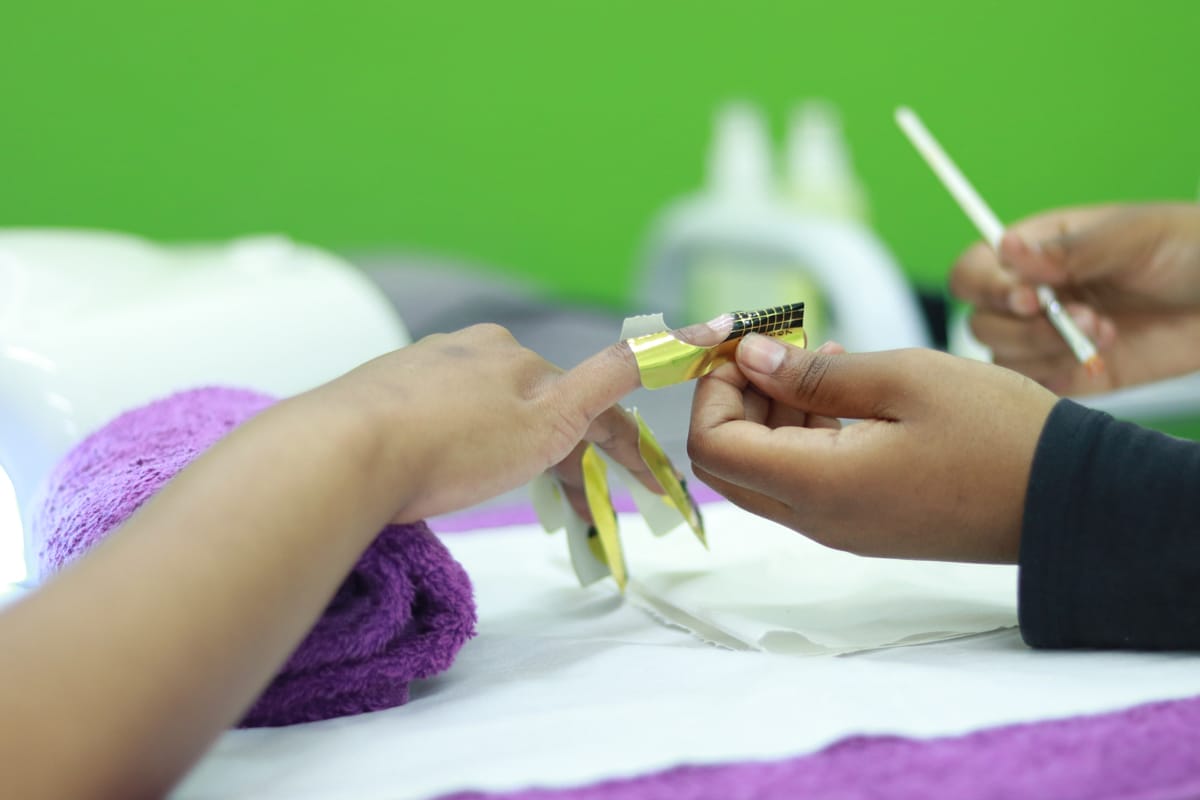 A close-up of a person receiving a manicure. One hand holds the other’s finger, applying a gold nail form for sculpting. A purple towel, white table, and green background are visible, with manicure tools blurred in the background.