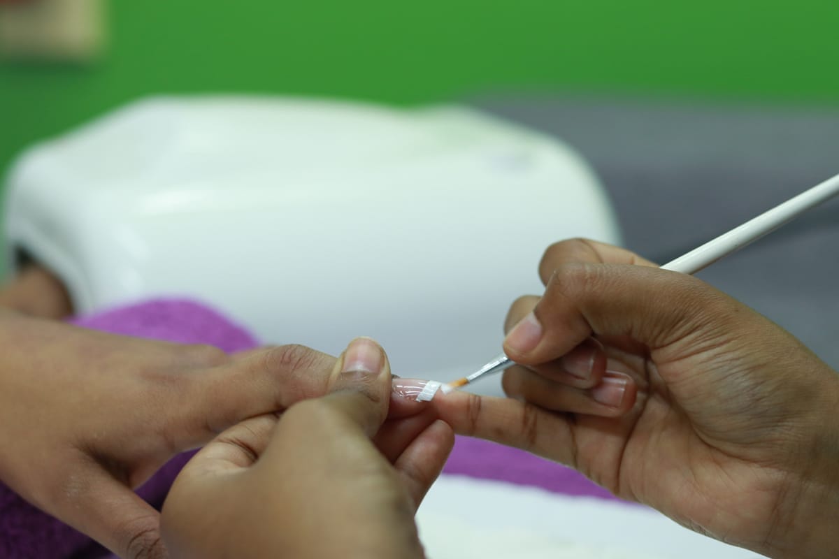 Close-up of hands during a manicure; one person paints clear varnish on another’s fingernail using a small brush. The background is blurred, with a green wall and a white UV nail lamp partially visible. A purple towel is under the person's hand.