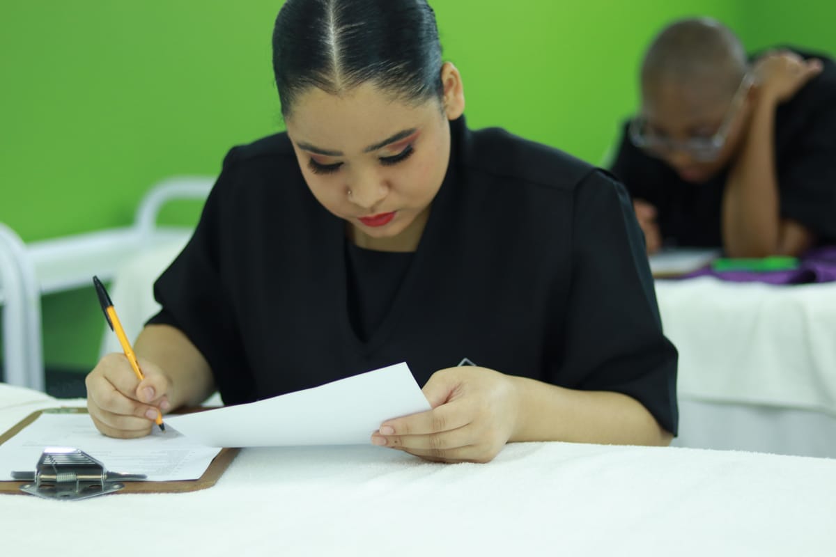 A woman with dark hair tied back, wearing black, writes on paper with a pen, focused on her work at a table. Another person sits behind her, also working, in a room with bright green walls and white tables.