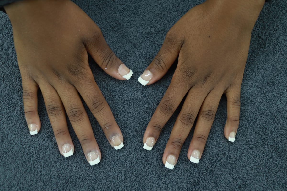 Two hands with medium brown skin rest on a dark textured surface. The nails have a French manicure style, with white tips and a natural pink base. The fingers are spread slightly apart, and the nails are neatly shaped and glossy.