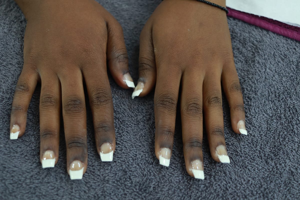 Two hands with medium brown skin rest on a grey towel. The nails are manicured with a classic French tip style—white tips and a natural pink base. The person wears a black string bracelet on their right wrist. The fingers are spread slightly apart.