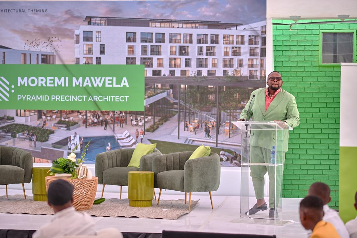 A man in a light green suit and sunglasses stands at a clear lectern, speaking to an audience. Behind him is a large poster showing a modern building and the text 