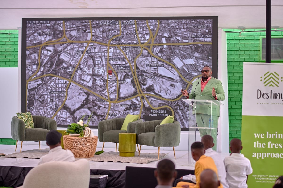 A man in a green suit speaks at a lectern on stage, with a large map projected behind him. The map shows city streets and neighbourhoods. People, including children in white and yellow, sit facing the stage. Green chairs and a table are also on stage.