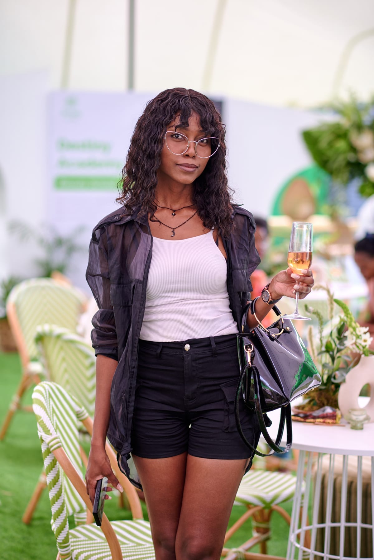 A young woman with curly hair and glasses stands indoors, holding a glass of champagne. She wears a sheer black shirt over a white vest, black shorts, and carries a shiny black handbag. The background features green chairs, white tables, and decorative plants.
