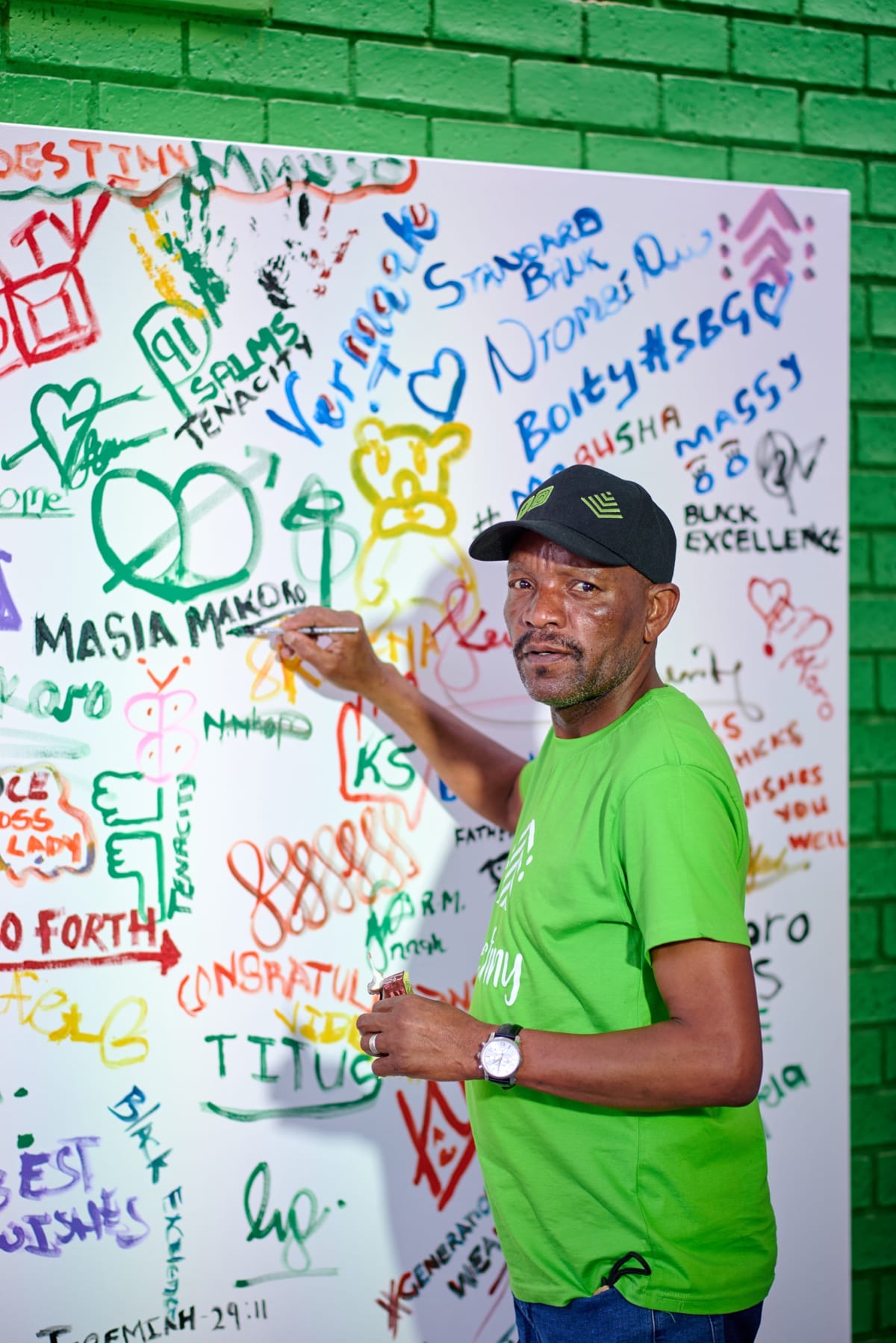 A man in a green T-shirt and cap stands in front of a graffiti-covered whiteboard and green brick wall, holding a marker and pointing at the board. The board displays colourful drawings, signatures, and messages in various handwriting styles.