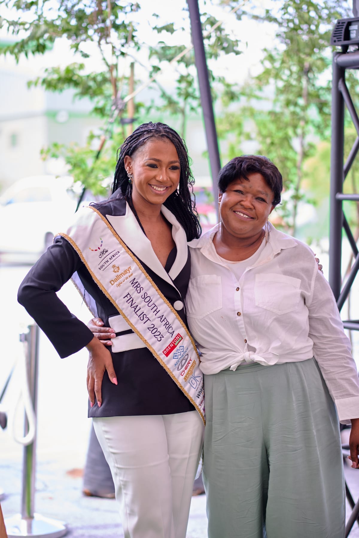 Two women stand side by side, smiling. The woman on the left wears a black and white suit with a “Miss South Africa Finalist 2023” sash. The woman on the right wears a white blouse and light green trousers. Outdoor setting with blurred trees in the background.