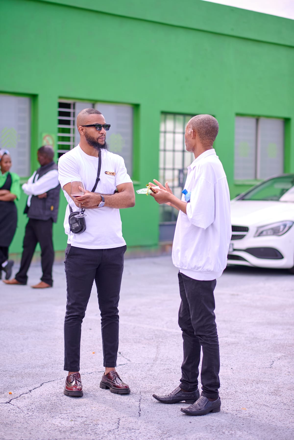 Two men stand and talk in a car park beside a white car and a green building. One man, wearing sunglasses and a camera across his shoulder, listens with arms folded. The other gestures while speaking. Two women and a man stand in the background near the building.