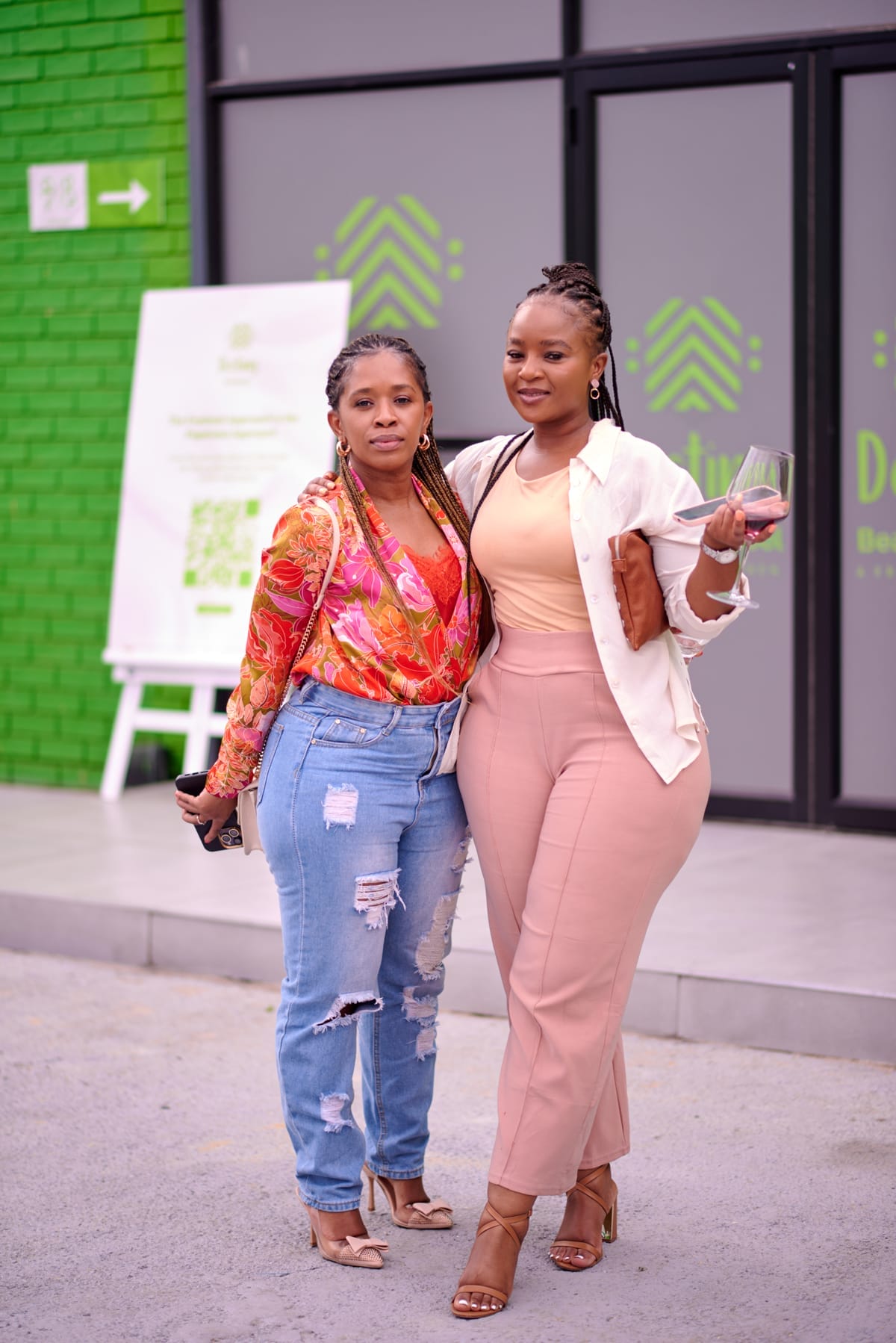 Two women pose outdoors in front of a green brick wall and glass doors. One wears a bright floral top with ripped jeans and sandals; the other wears a beige top and trousers with a white shirt, holding a drink. Both have plaited hair and smile at the camera.