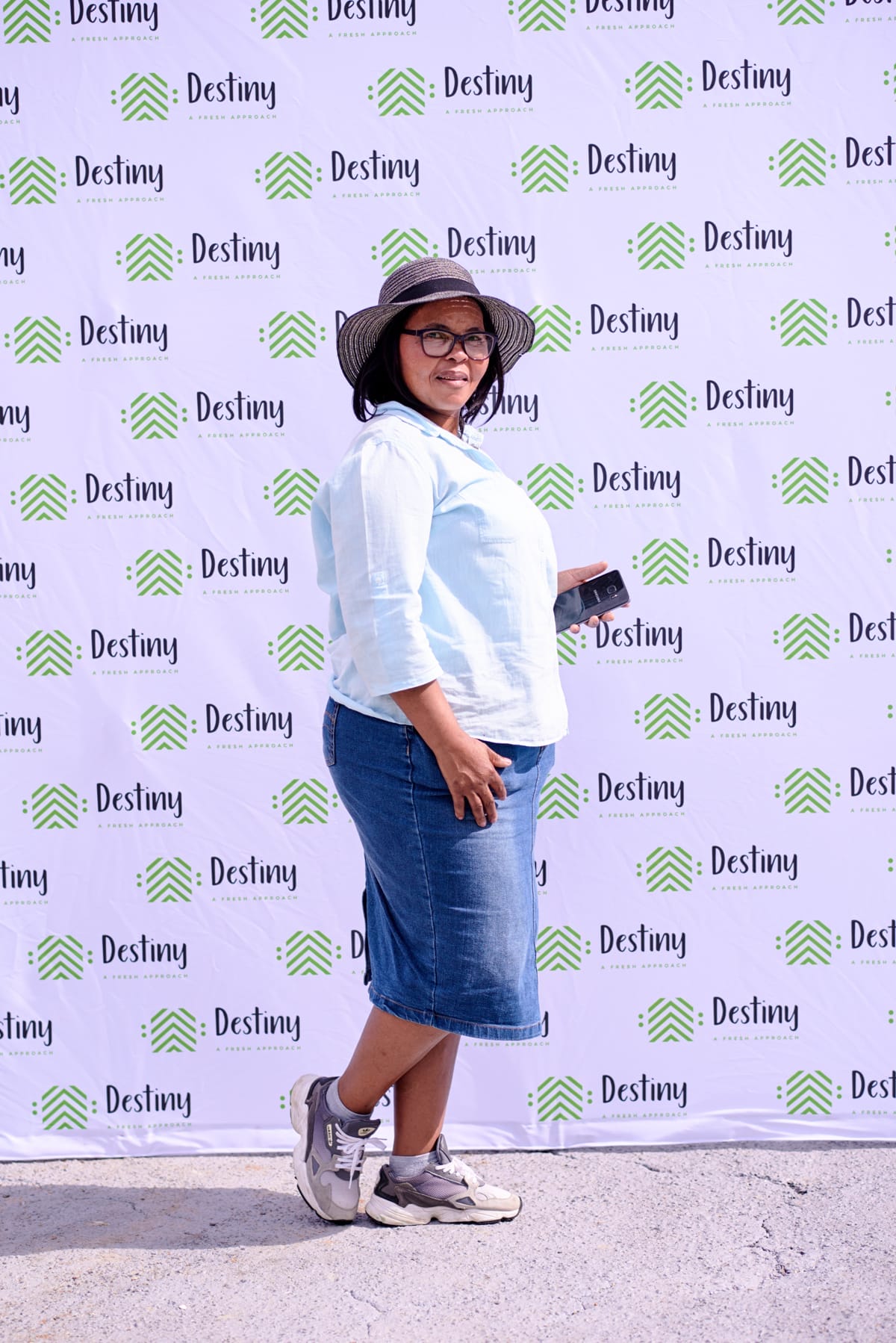 A woman in a light blue shirt, denim skirt, trainers, and a sunhat stands sideways, smiling and holding a mobile phone. She is in front of a backdrop with green and black 