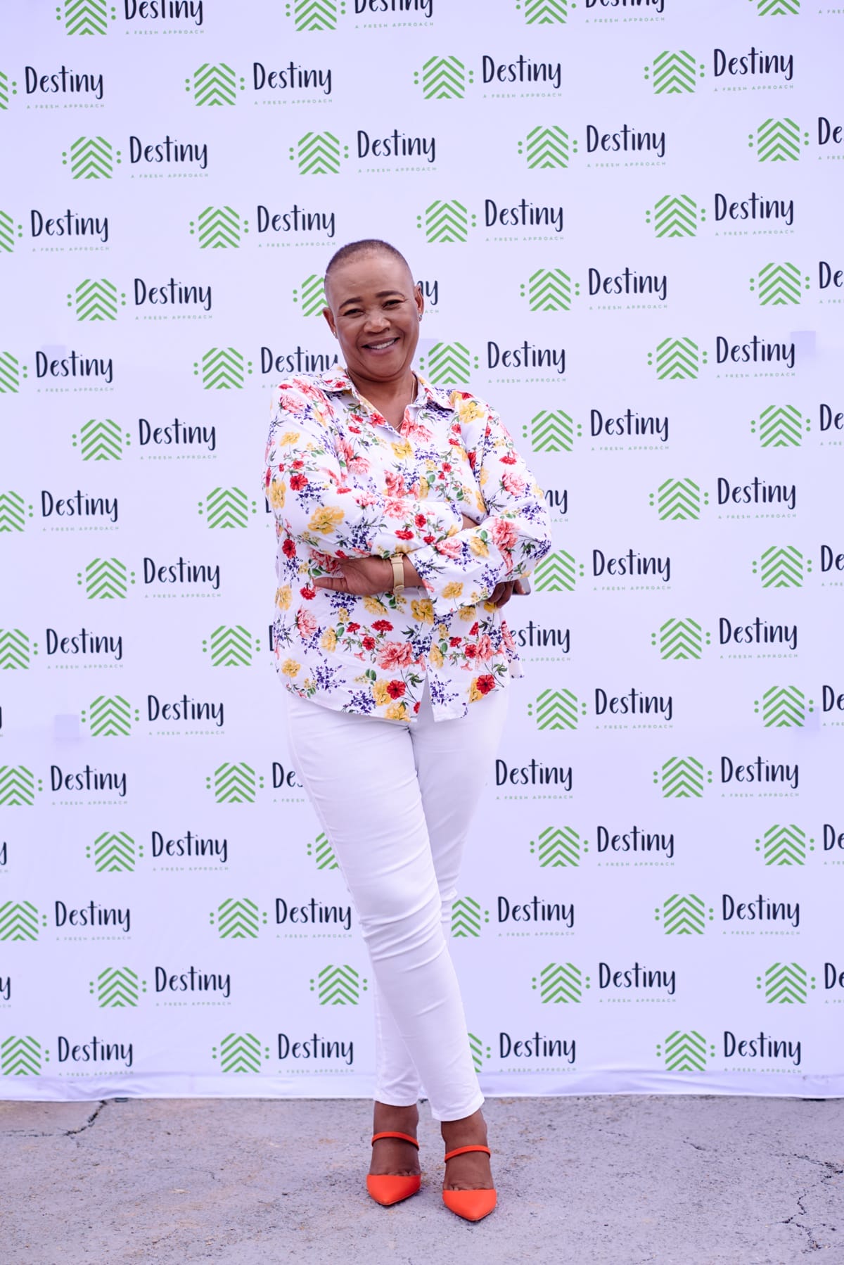 A smiling person with short hair stands confidently with arms crossed, wearing a floral shirt, white trousers, and red shoes. The background features a white step-and-repeat banner with green logos and the word “Destiny” repeated in black text.