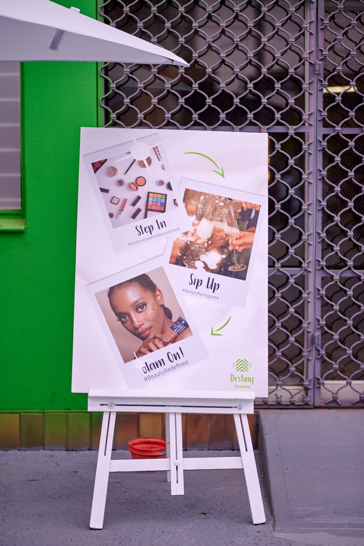 A poster on an easel stands outside near a green wall and a metal gate. It features make-up items, a woman’s portrait, and people clinking glasses, with the phrases: “Step In”, “Sip Up”, and “Glam On!” The Destiny Foundation logo appears at the bottom.