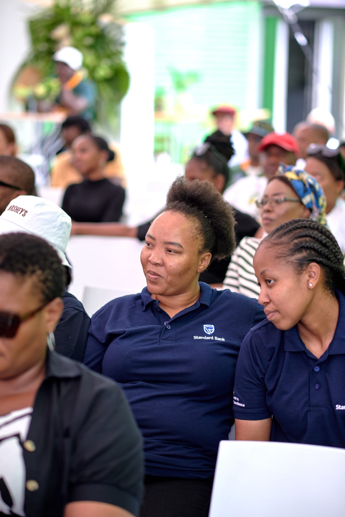 A group of people sit closely together at an indoor event. Two women in the foreground wear navy blue Standard Bank shirts and are looking to the side, smiling. Other attendees, some in colourful headscarves, are seated in rows behind them. The background is softly blurred.