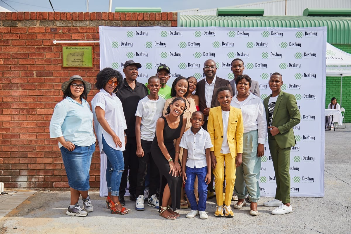 A group of 14 smiling people, including adults and children, pose together outdoors in front of a red brick wall and a step-and-repeat banner with “Destiny” logos. Some wear casual clothes, while others are dressed formally. The background includes a green and white building.
