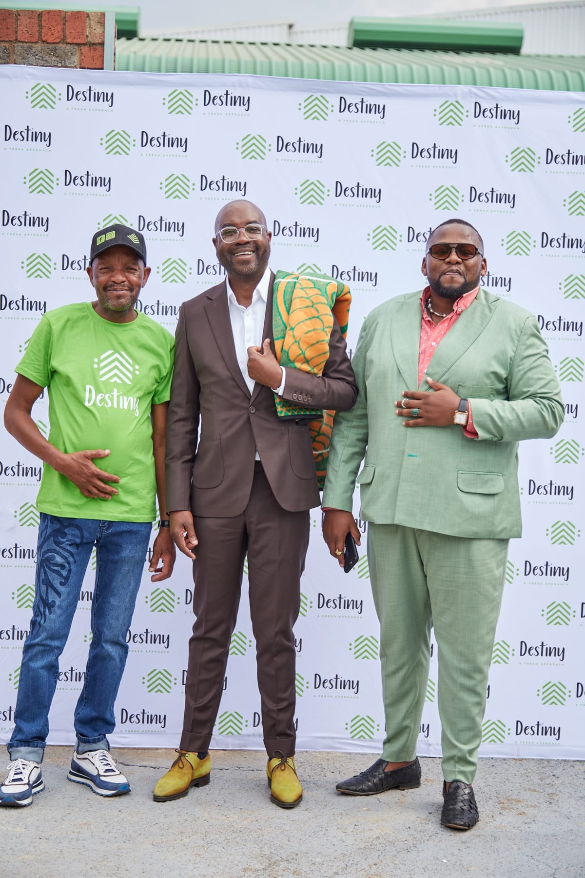 Three men stand smiling in front of a white “Destiny” step-and-repeat banner. One wears a green Destiny T-shirt, jeans, and a cap. The middle man wears a brown suit and holds a colourful patterned fabric. The third man wears a light green suit and sunglasses.