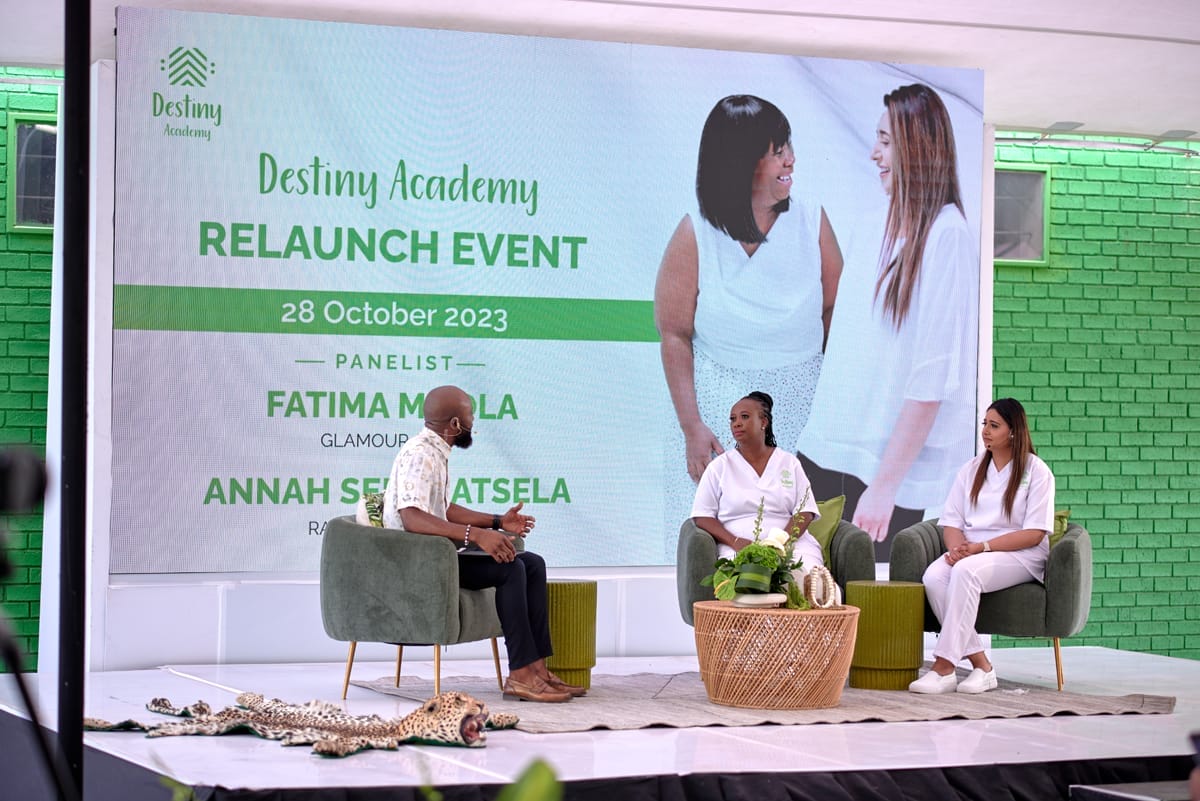 Three people sit on a stage in front of a large screen reading “Destiny Academy Relaunch Event, 28 October 2023,” with panellist names. Two women in white chat with a man in brown; the backdrop and walls are green. A plant decorates the table beside them.