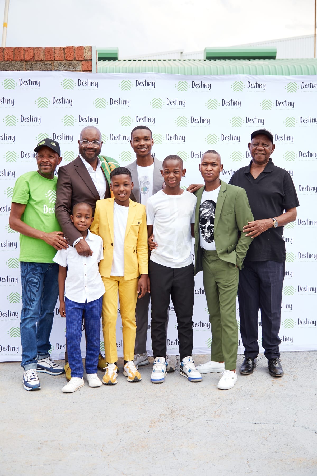 Eight men and boys stand together smiling in front of a white Destiny-branded backdrop. They wear a mix of formal and casual outfits in green, yellow, black, and white tones, suggesting a celebratory or formal event. Everyone faces the camera, posing closely.