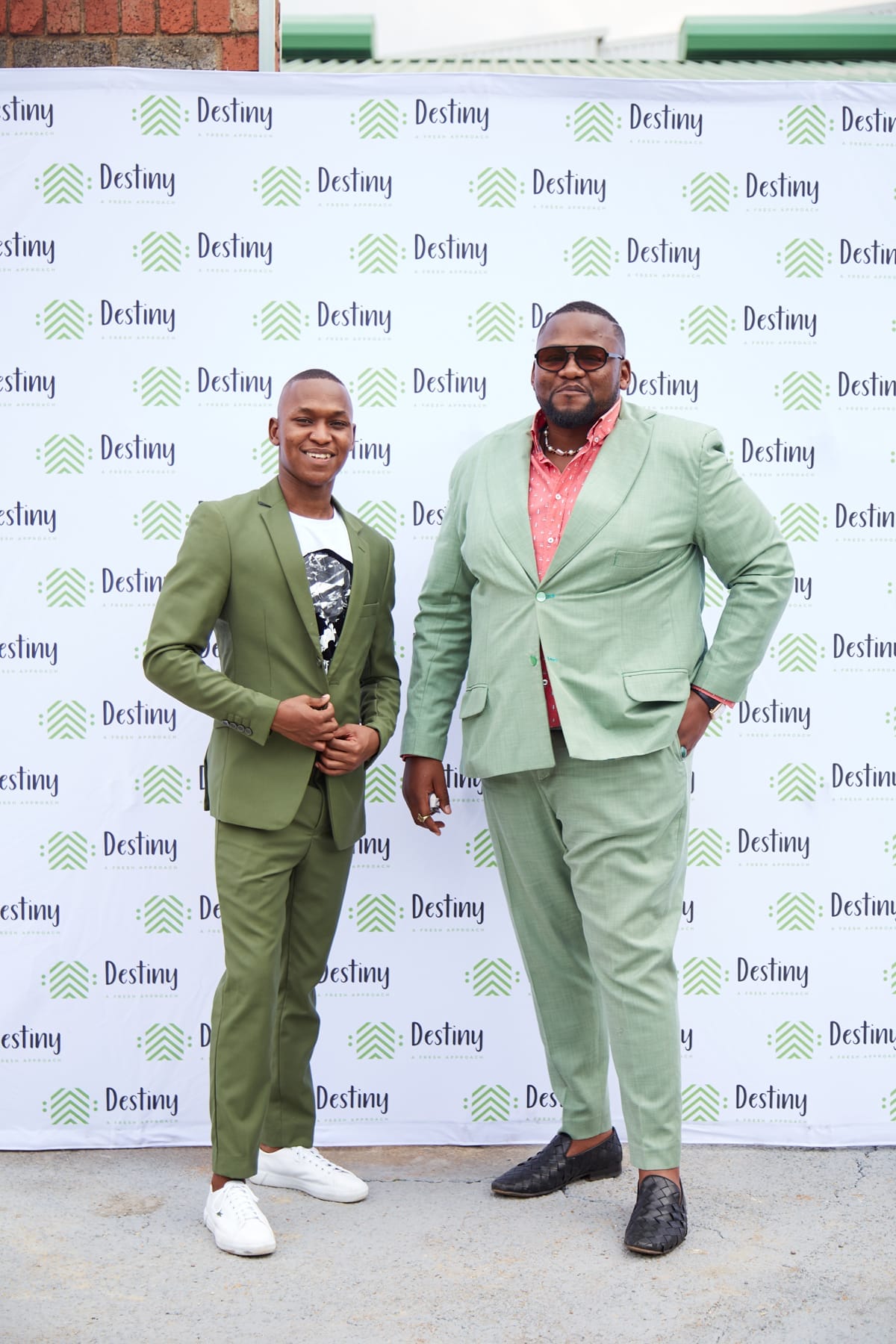 Two men pose in stylish green suits in front of a white backdrop with green and black “Destiny” logos. The man on the left is shorter, smiling, wears trainers, and a graphic t-shirt. The taller man has sunglasses and a red shirt, and wears smart shoes.