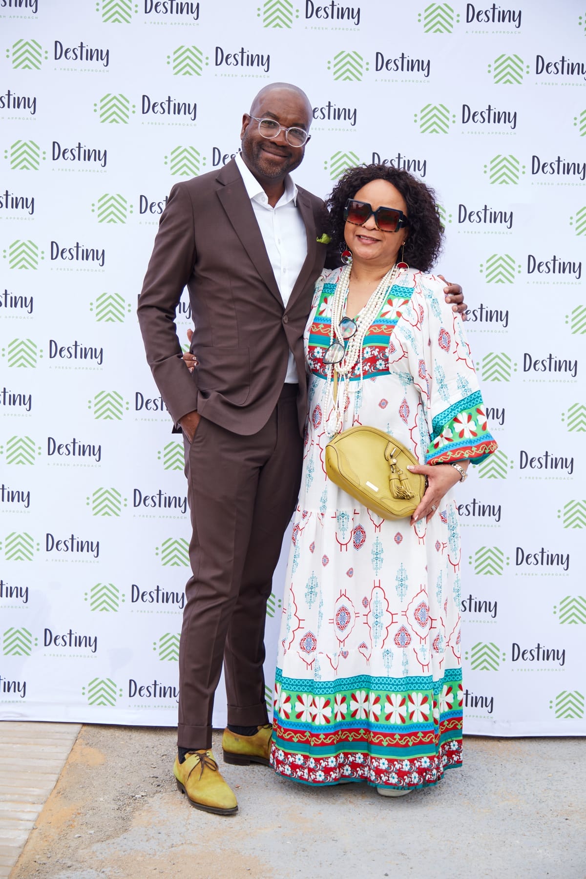 A man in a brown suit and yellow shoes stands with a woman in a colourful patterned dress, sunglasses, and a yellow handbag. They smile and pose together in front of a white backdrop with “Destiny” and green logo patterns.