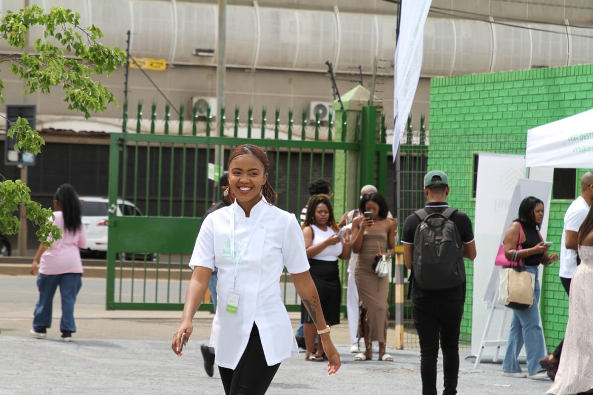 A smiling woman in a white top walks outdoors towards the camera. Behind her, several people stand near a green brick wall and an iron gate. Cars and urban buildings are visible in the background under a cloudy sky.
