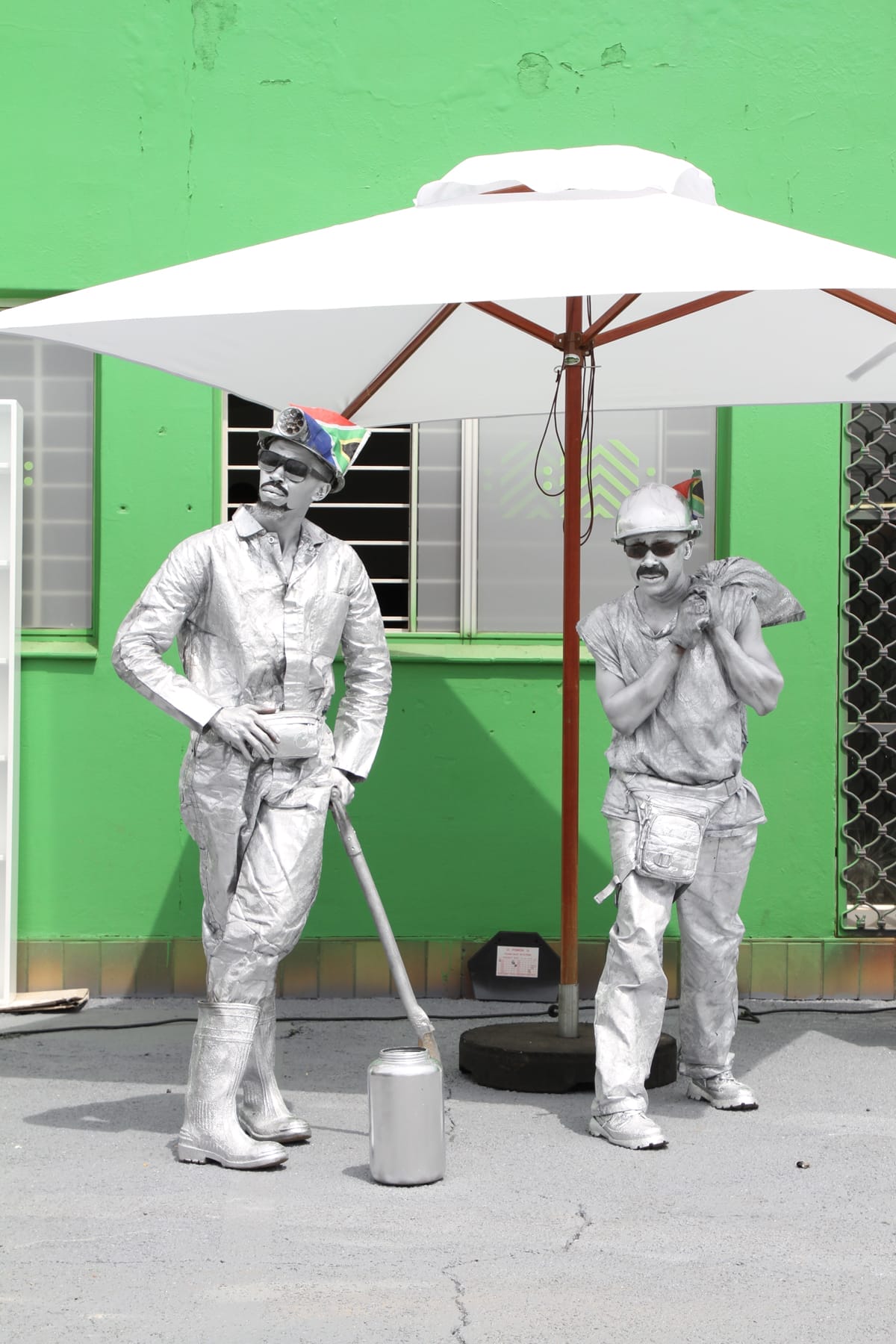 Two men dressed as silver-painted statues stand under a large white umbrella. Both wear silver outfits, hats, and sunglasses; one holds a sledgehammer, the other has a pickaxe over his shoulder. The background is a bright green wall with windows.