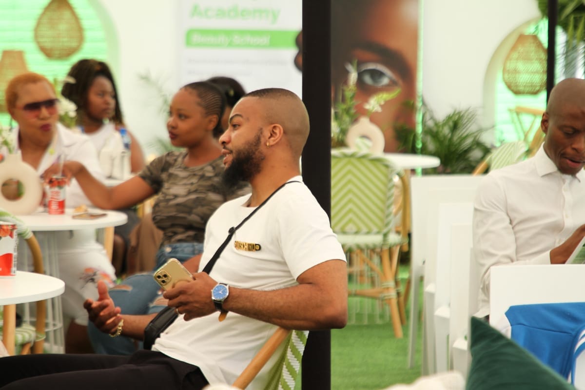 A bearded man in a white T-shirt sits talking at a table in a bright, modern café. Other people sit nearby, chatting and holding drinks. Green and white décor, plants, and a “Beauty School” banner are visible in the background, creating a lively atmosphere.