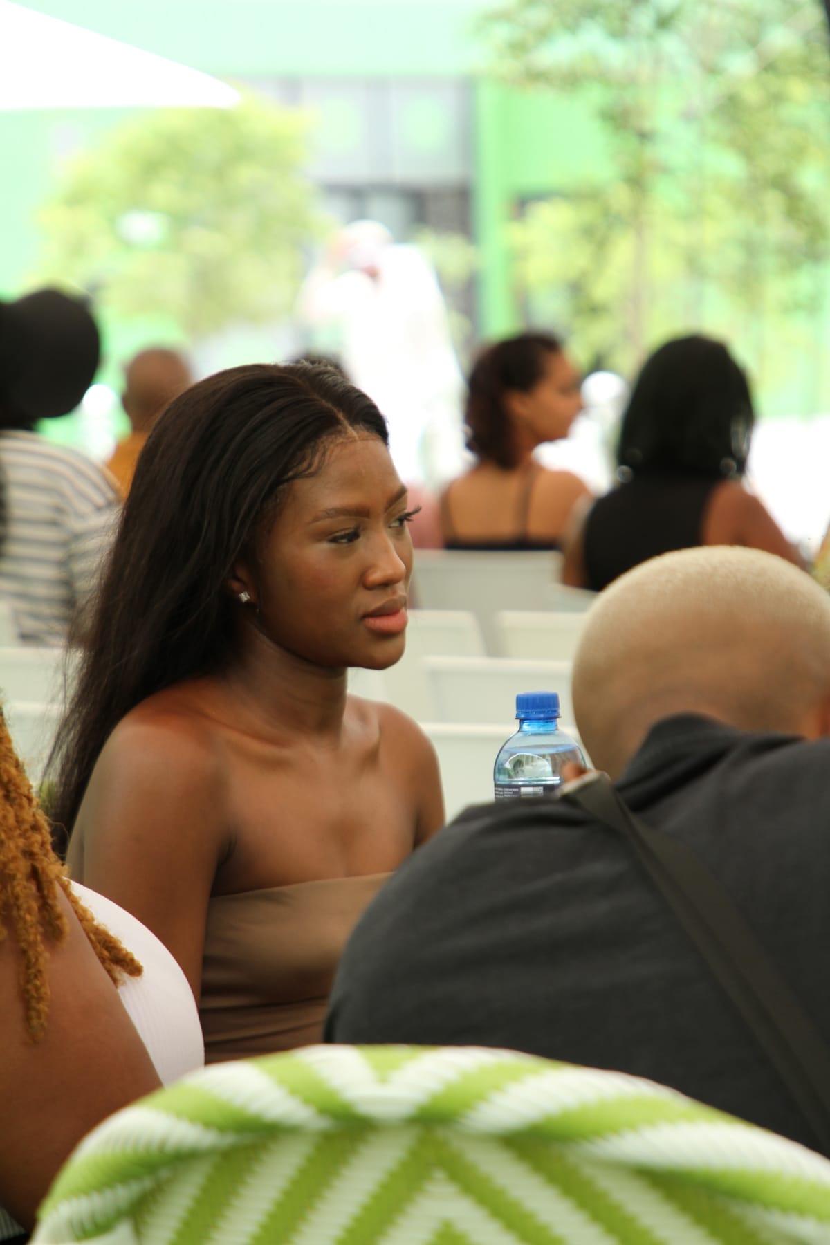A young woman with long dark hair and a brown strapless top sits at an outdoor gathering, looking engaged. Other people, including a bald person holding a water bottle, sit nearby. The background is blurred, with green tones and trees visible.