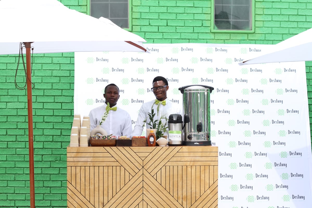 Two men in white shirts and green bow ties stand behind a wooden coffee stall with cups, snacks, and a large coffee urn. White umbrellas shade the area. Behind them is a bright green brick wall and a Destiny-branded backdrop.
