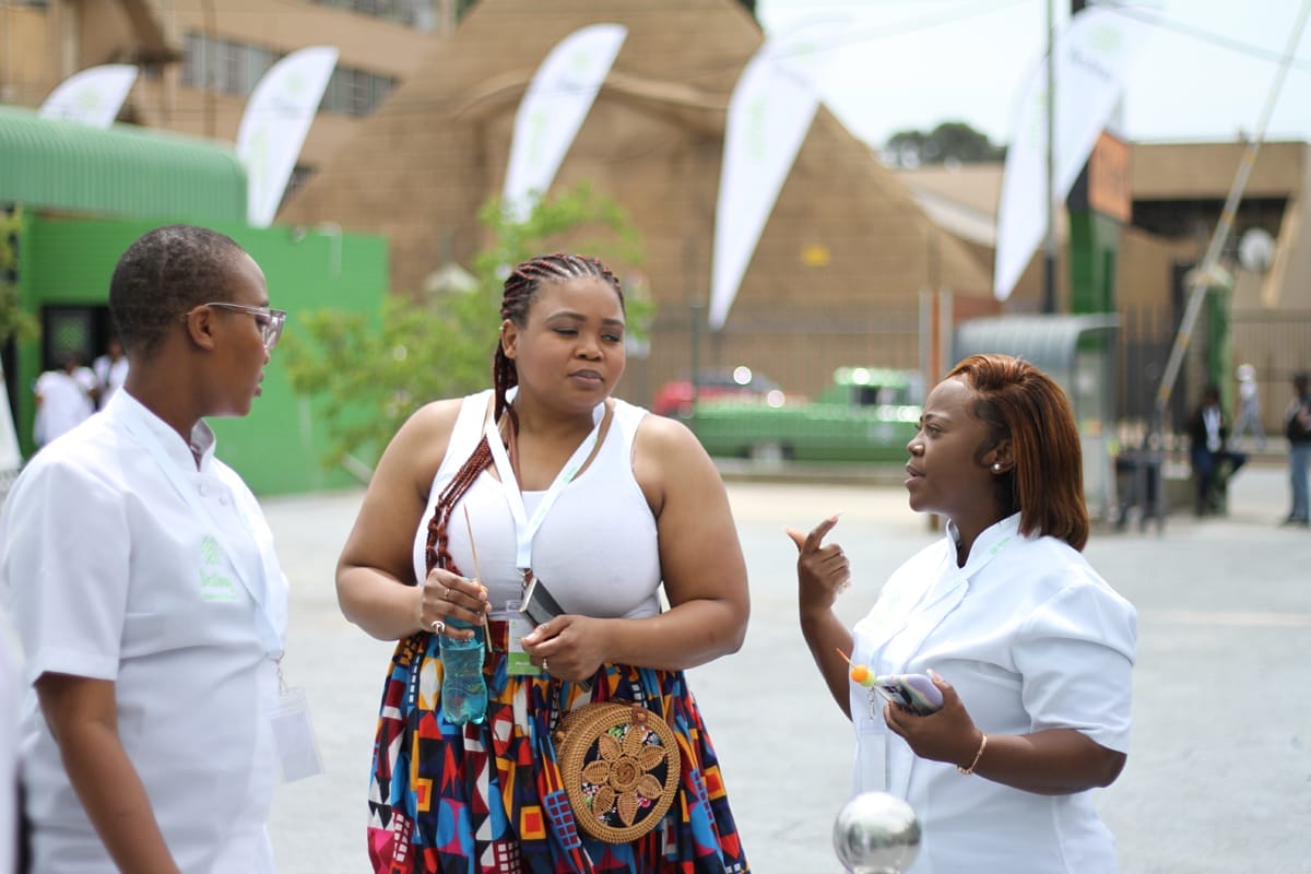Three women stand outside talking. Two wear white tops with name badges; one holds a water bottle and has a colourful skirt with a round woven bag. They appear engaged in conversation, with blurred white flags and buildings in the background.
