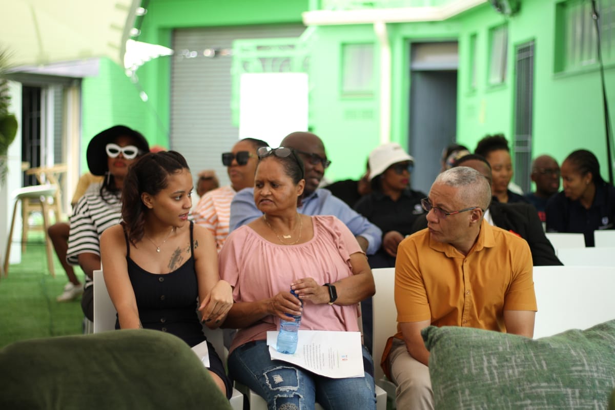 A diverse group of people sit in rows at an indoor event with bright green walls. Three people in front—a young woman, an older woman holding water, and a man in an orange shirt—converse, while others in the background watch or chat.