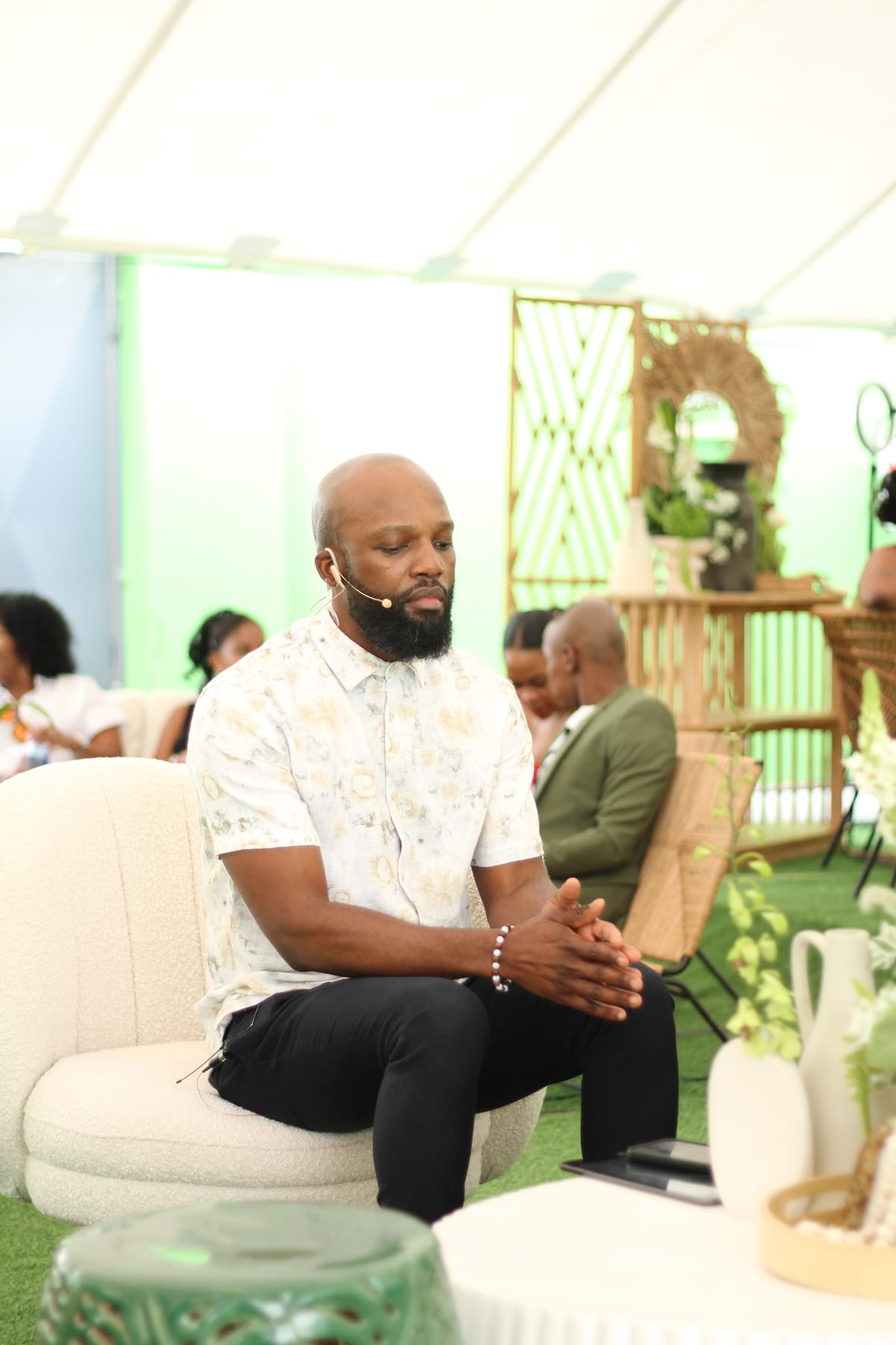 A man with a beard, wearing a white patterned shirt and black trousers, sits on a round chair with his hands clasped. He wears a headset microphone and has his eyes closed, as if meditating. The setting is bright, relaxed, and modern, with plants and people in the background.