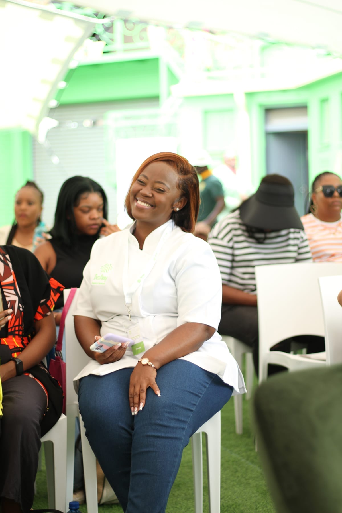 A woman in a white shirt and blue jeans sits smiling on a white chair at an outdoor event. She holds her mobile and a notepad. Other people sit and listen around her, with green structures and natural light in the background.