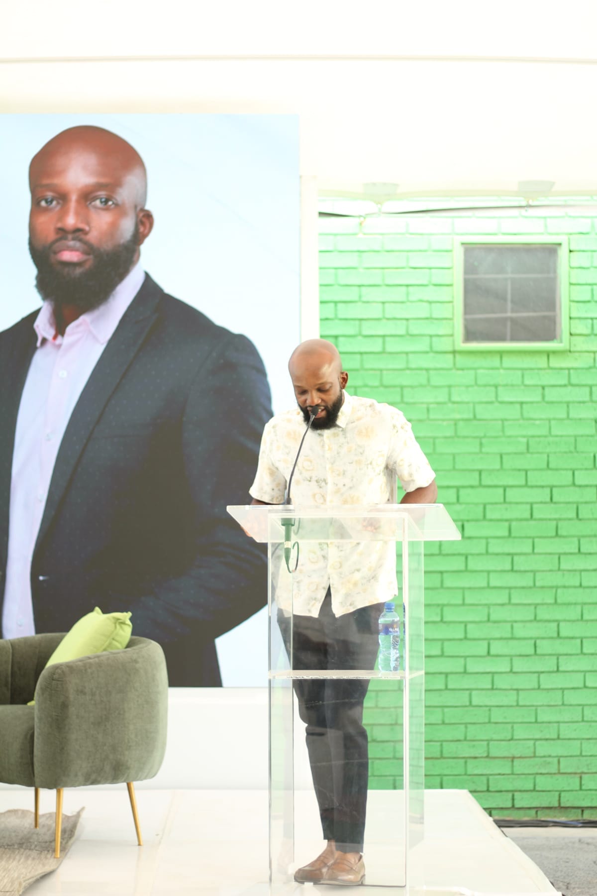 A bald man with a beard stands reading at a clear lectern. Behind him is a large portrait of himself in a suit on a green brick wall. A green armchair with a cushion sits beside him. The setting appears modern and well-lit.