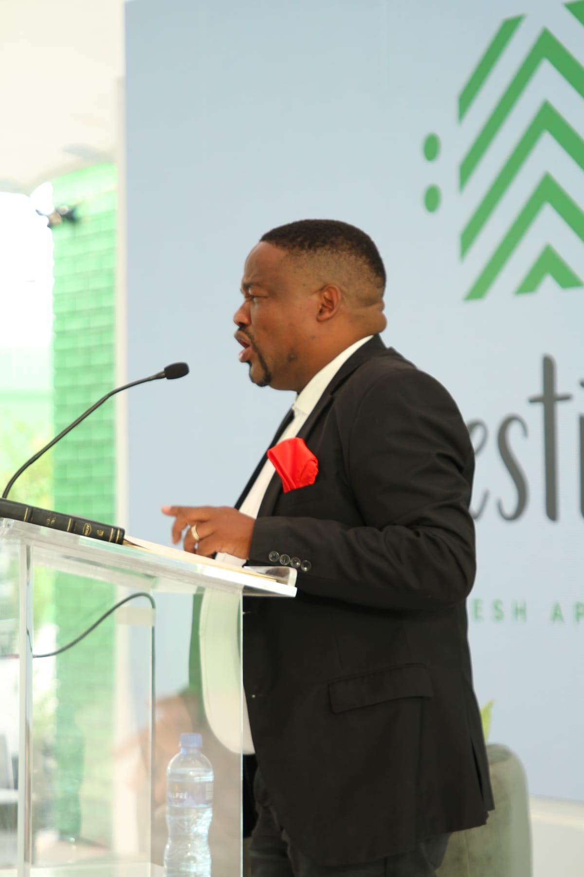 A man in a black suit with a red pocket square speaks passionately at a clear lectern with a microphone. A water bottle and book are on the lectern. Behind him is a blue wall with a green geometric logo and partial text visible.