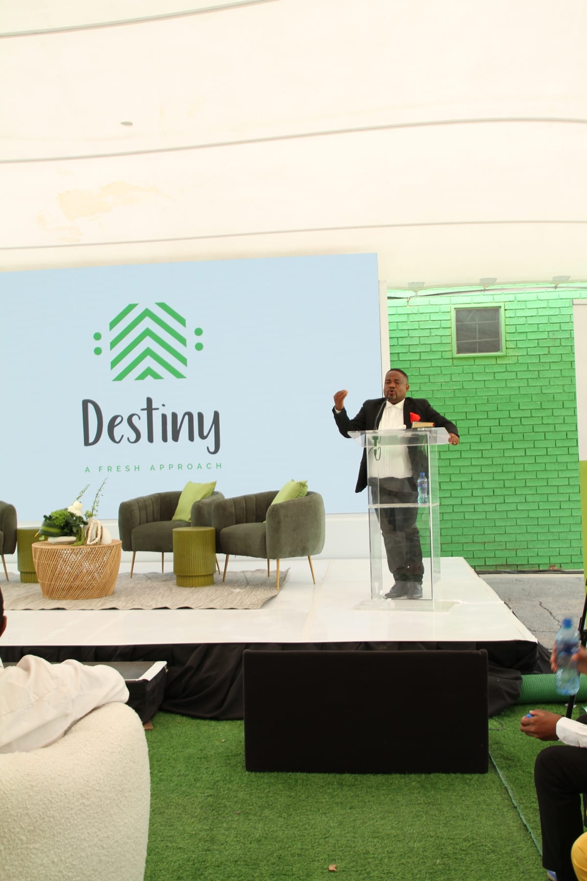 A man in a black suit speaks at a clear lectern on a white stage with green chairs and plants. Behind him is a large screen displaying the word 