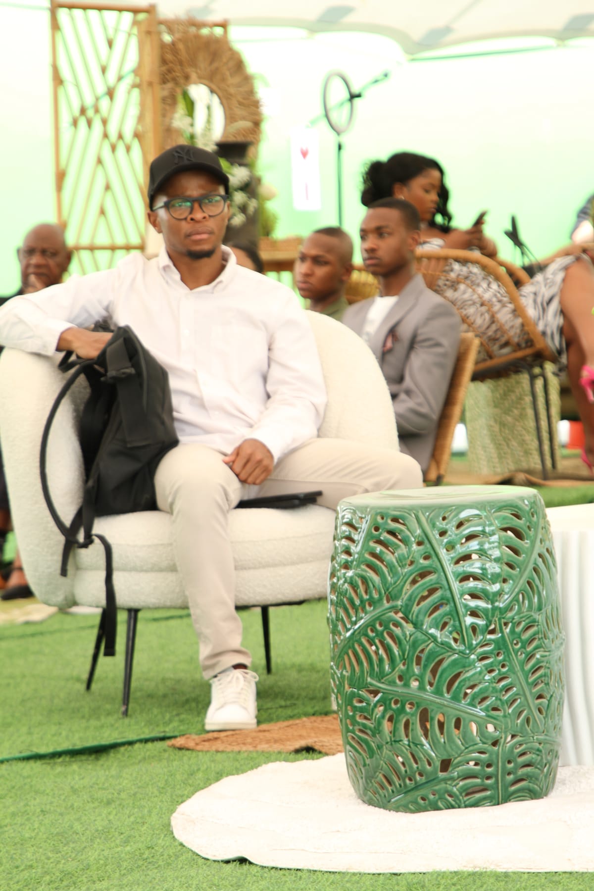 A young man wearing glasses, a black cap, white shirt, and beige trousers sits on a modern white chair holding a rucksack. In front of him is a green ceramic stool with a leaf pattern. Several people are seated in the background on wicker chairs. The setting appears casual and indoors.