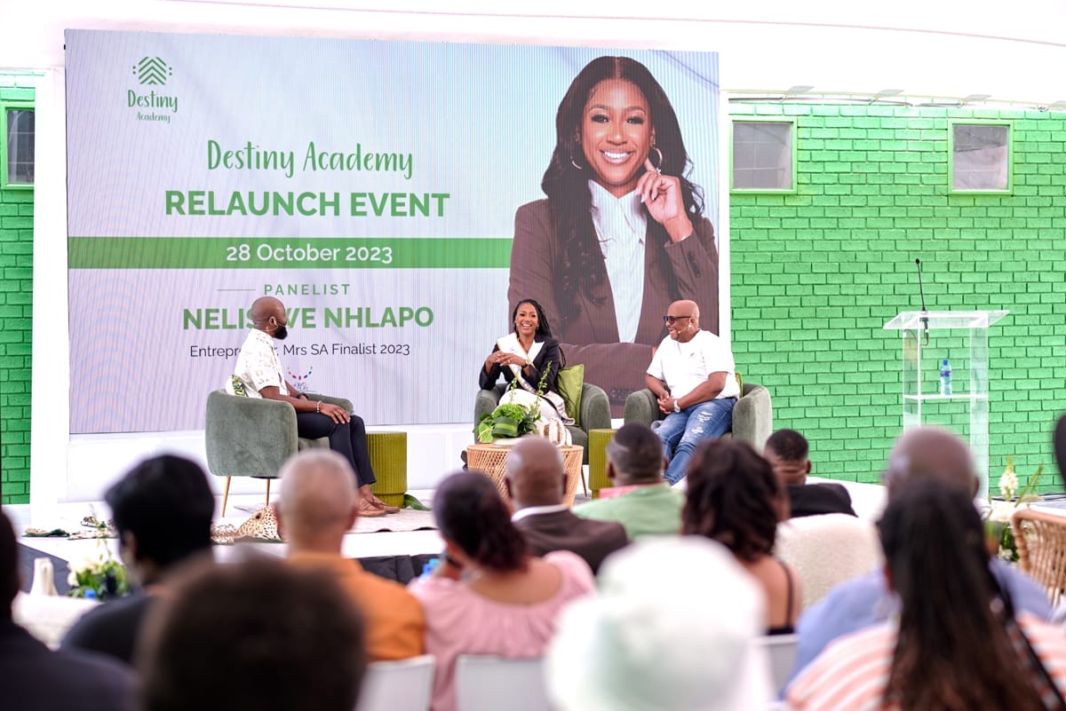 Three people sit on stage in front of an audience at the Destiny Academy Relaunch Event. Behind them is a green and white banner with a photo of a smiling woman and event details. The setting is bright, with green brick walls and floral décor.