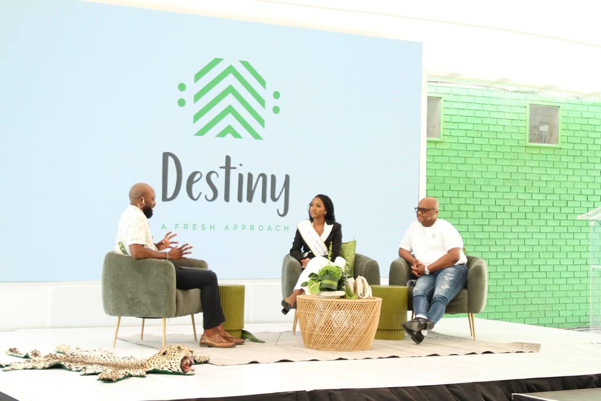 Three people sit in modern chairs on a stage, engaged in discussion. Behind them is a large screen with 