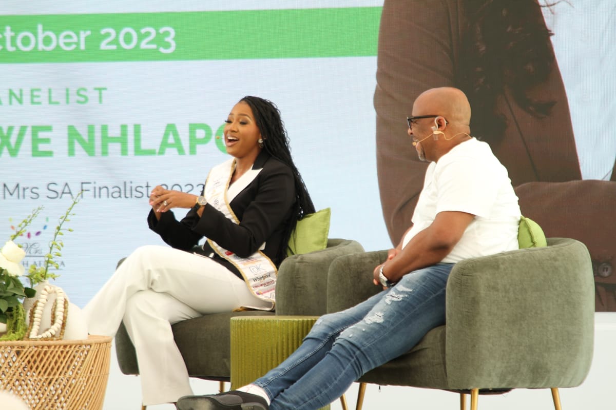 Two people sit on stage in green armchairs, having a conversation. The woman on the left wears a sash and a black blazer, gesturing as she speaks. The man on the right wears glasses, a mic headset, a white T-shirt, and jeans. A plant decorates the foreground.