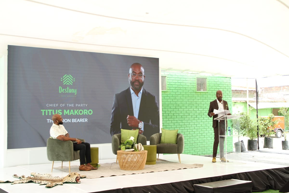 A man in a suit speaks at a lectern on a stage with green decor. Behind him, a large screen displays his photo, the Destiny party logo, and text: 