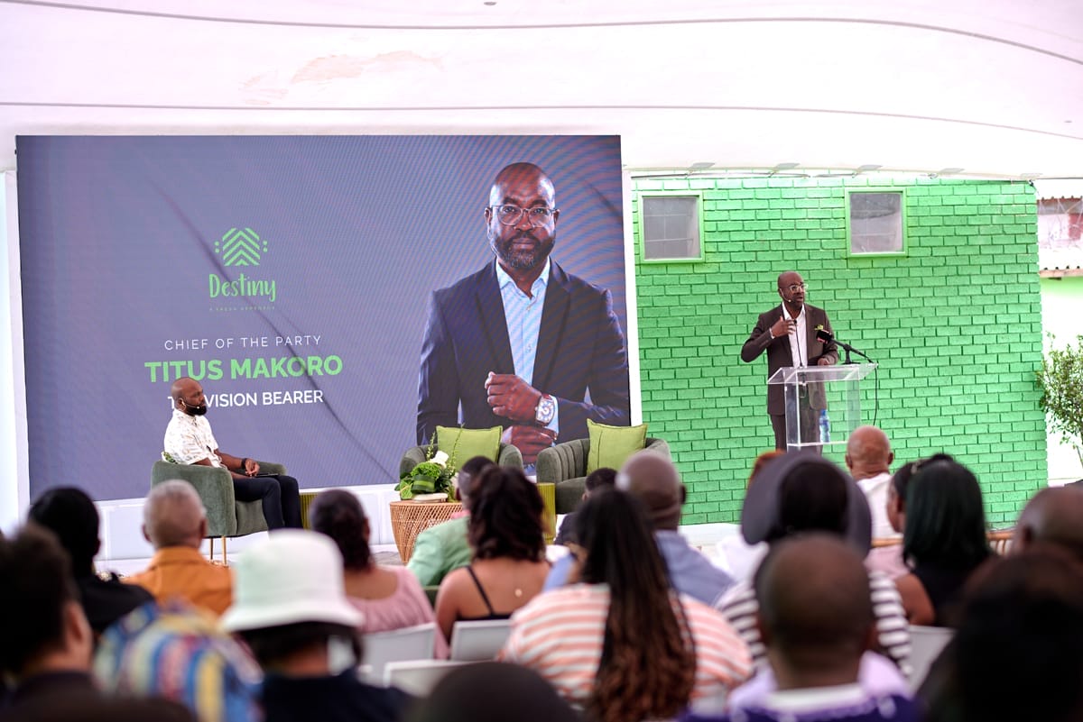 A man speaks at a clear lectern to an audience indoors. Beside him is a large screen displaying his photo and text: “Destiny, Chief of the Party, Titus Makoro, Vision Bearer.” Another man sits onstage. The setting features green décor and seating.