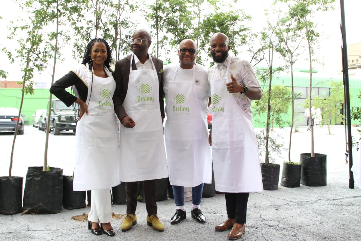 Four people stand side by side outdoors, smiling at the camera. They wear white aprons with a green “Deslay” logo, in front of young trees in black pots. Three people have their arms round each other; one gives a thumbs-up. Cars and a green building are visible in the background.