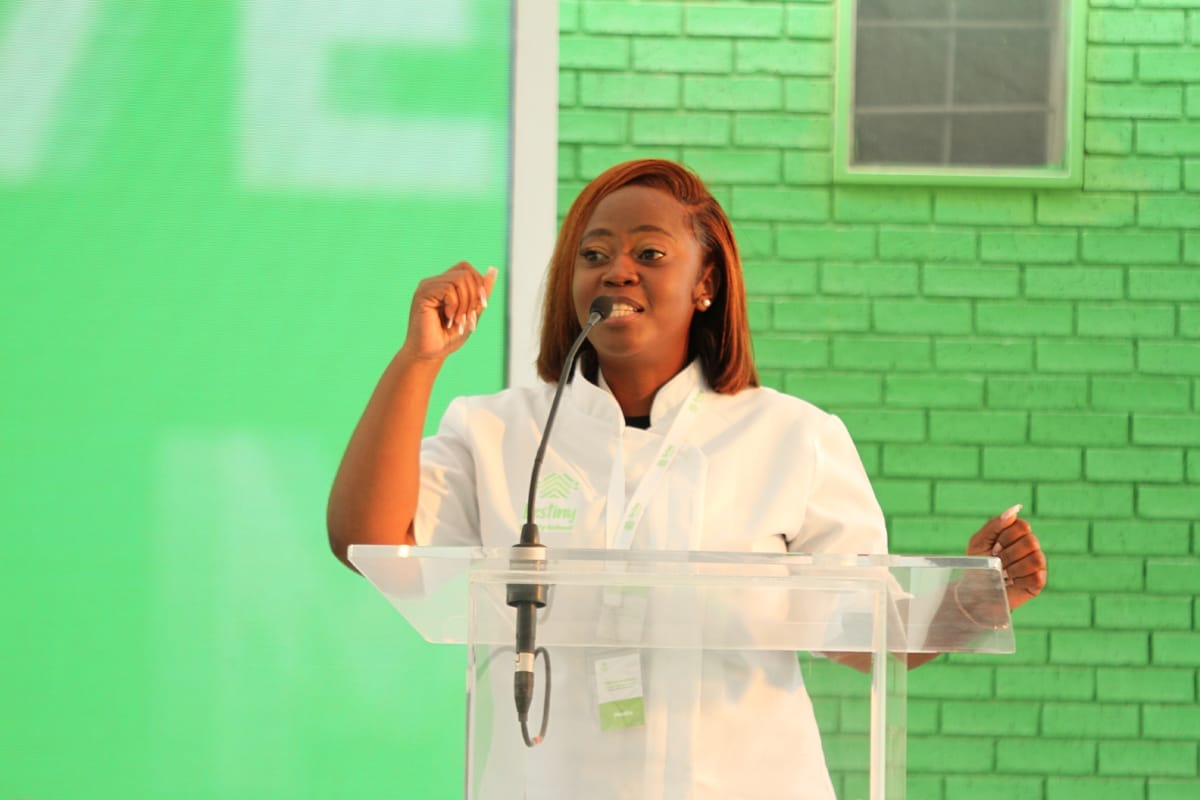 A woman with straight brown hair speaks passionately at a clear lectern, wearing a white shirt and conference badge. She gestures with one hand raised. The background features a green brick wall and a large green screen.