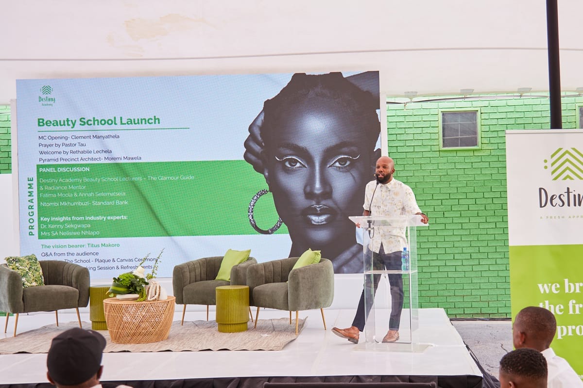 A man stands at a lectern on a stage, speaking at the Beauty School Launch event. Behind him is a large illustration of a woman's face, a green programme schedule, and a green wall. Modern chairs and a small table with decor are on the stage.