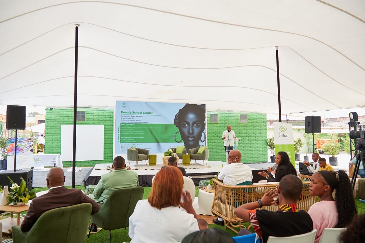 A diverse audience sits on green and wicker chairs under a white canopy, facing a stage with a large screen displaying a woman's face and presentation slides. A person stands at a lectern near green brick walls, and plants decorate the event space.